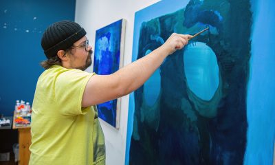 photo of an artist standing, arm extending brushing blue paint on a canvas with many abstract shapes in shades of blue.