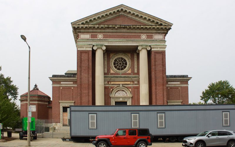 A large Italian Renaissance Revival church with brick exterior and two terra cotta columns beside the front entrance. A large gray construction trailer and two cars are parked before the church.