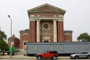 facade of a large Italian Renaissance Revival church with brick exterior and two terra cotta columns beside the front entrance. A large gray construction trailer and two cars are parked in front of the church.