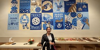 photo of the inside of a restaurant, a woman stands in front of a hand painted grid of images in various shades of blue.