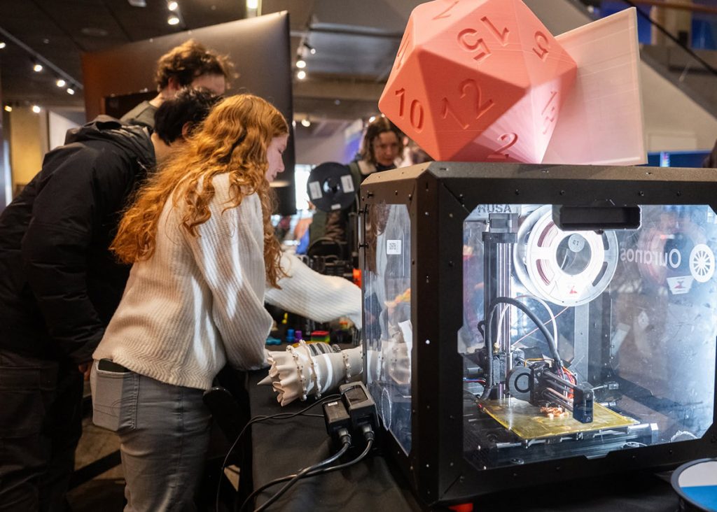 People gather around an enclosed 3D printer topped with a large translucent pink die in a maker space inside the Museum of Science.