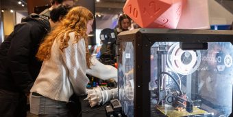 People gather around an enclosed 3D printer topped with a large translucent pink die in a maker space inside the Museum of Science.