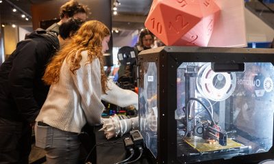 People gather around an enclosed 3D printer topped with a large translucent pink die in a maker space inside the Museum of Science.